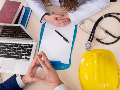 Biurko z laptopem, długopisem, kartką, stetoskopem, kaskiem i ręce lekarki i pacjenta