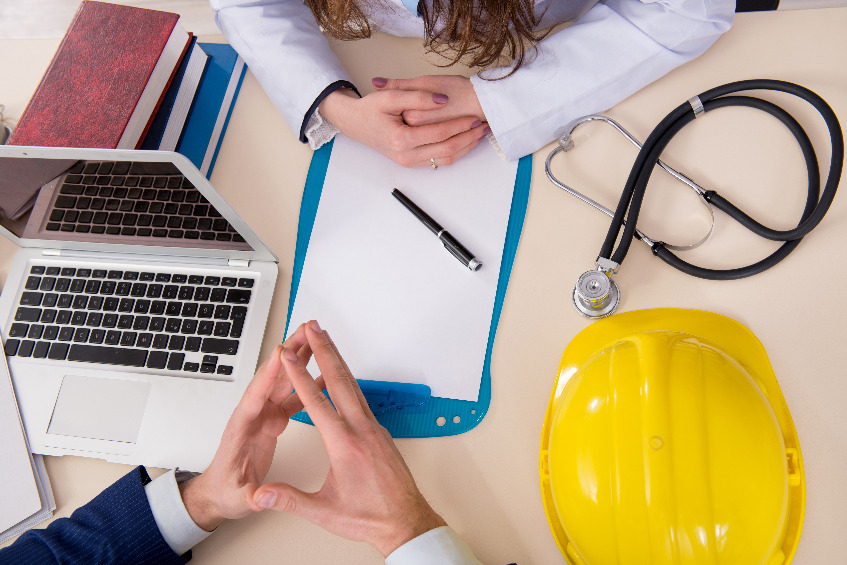 Biurko z laptopem, długopisem, kartką, stetoskopem, kaskiem i ręce lekarki i pacjenta