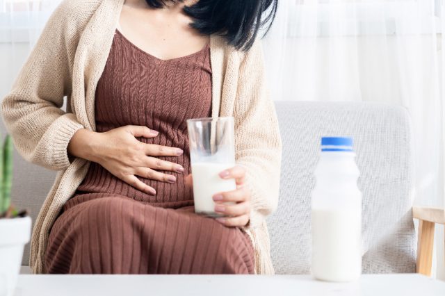 Kobieta trzymająca się za brzuch jedną ręką, a w drugiej trzymająca szklankę z mlekiem